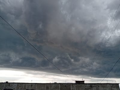 The image depicts a cloudy sky over a building in Kyshtym, Russia. Dark, heavy storm clouds dominate the upper portion of the frame, with lighter clouds and a brighter sky visible near the horizon. Multiple power lines crisscross the sky. Below the clouds, the top of a concrete building is visible, characterized by a textured surface and a railing along its edge. Small structures, possibly vents or chimneys, are present on the roof. The overall atmosphere suggests an impending storm.