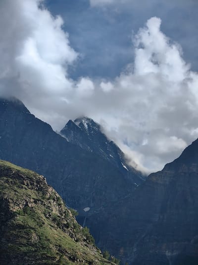 The image captures a majestic mountain landscape under a dynamic sky. In the foreground, a steep, rocky slope is covered with patches of green grass and scattered trees. Behind this, dark, imposing mountains rise dramatically, their rugged surfaces textured with layers of rock and debris. Snow-capped peaks are visible in the distance, partially obscured by swirling clouds. The sky is a mix of deep blue and bright white, with dramatic formations of cumulus clouds casting shadows on the mountains below. The overall scene evokes a sense of grandeur and the raw beauty of nature. This image is located in Tiling, India.