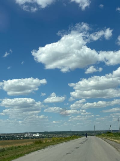 The media file depicts a landscape with a clear blue sky filled with scattered white clouds. A paved road curves to the right, bordered by grassy fields. In the distance, a town or city is visible with various buildings. A single motorcycle with its headlights on is seen on the road, heading away from the viewer. Several utility poles are scattered along the side of the road. The scene suggests a sunny day, likely daytime, and the open road hints at a journey or travel. The overall impression is one of a rural or semi-rural setting on a clear day.