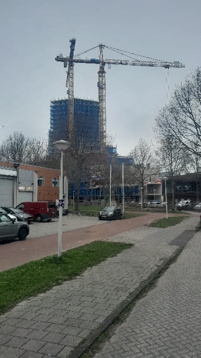 The image displays an urban environment in Amsterdam-Duivendrecht, Netherlands, under an overcast, light grey sky. In the background, a multi-story building under construction is enveloped by blue scaffolding and safety netting. Two large tower cranes, one with a dark blue upper section and another yellow/orange, are positioned above and next to the building. The midground includes several bare deciduous trees, patches of green grass, and various finished commercial or residential buildings. A paved parking area to the left contains parked vehicles, including a dark grey crossover SUV, a dark van, and a red cargo van with an attached trailer. A prominent white lamppost stands in the mid-left, next to a blue parking sign. A red-paved path runs horizontally in the midground, separated from a grey-paved sidewalk in the foreground by a strip of grass. The sidewalk consists of rectangular pavers, with a herringbone pattern visible closer to the bottom right. No individuals are present.