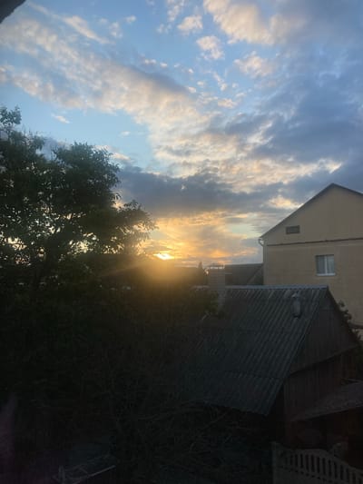 The image depicts a sunset scene in Матыкальскі сельскі Савет, Belarus, viewed from a slightly elevated perspective. The sky is filled with dramatic clouds, with the setting sun casting a warm, golden glow through the gaps. In the foreground, a large, dark tree with dense foliage dominates the left side of the frame, its branches silhouetted against the bright sky. To the right, two buildings are visible. The larger building is a light-colored, multi-story structure with visible windows and a downspout. In front of it, a smaller, darker building with a corrugated roof and a chimney is partially obscured by the tree. The overall impression is one of a rural or suburban setting at dusk. There are no people or animals visible in the image.