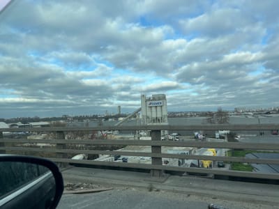 This image captures an industrial landscape in Gennevilliers, France, viewed from an elevated perspective, likely a bridge or overpass. The sky is partially cloudy with patches of blue visible through grey stratocumulus clouds.
In the immediate foreground, the top-left section of a dark vehicle's side mirror is visible, reflecting a distorted view of the road surface. Below this, the gritty, dark asphalt of a roadway extends, showing scattered debris and dirt. Horizontally across the lower half of the frame, a concrete bridge railing with two parallel beams and vertical supports separates the foreground from the midground.
The midground is dominated by an active industrial facility. A prominent CEMEX concrete batching plant stands centrally, featuring tall, light grey silos with the "CEMEX" logo prominently displayed. Surrounding the plant are paved areas and various industrial vehicles, including several cement mixer trucks (one yellow mixer clearly visible), other large commercial trucks, and smaller automobiles. A red bus-like vehicle and several dark-clad figures (people) are discernible in the far right midground, indicating human presence. The terrain is a mix of paved surfaces, dirt, and sparse, leafless trees.
The background presents an extensive urban and industrial panorama. Numerous low-rise buildings and warehouses stretch across the horizon. A distinct, tall, slender high-rise building (likely Tour Mozart) is visible in the distant background, signifying a more developed urban area beyond the immediate industrial zone. No specific actions or interactions are discernible due to the distance, beyond the general operation of an industrial site.