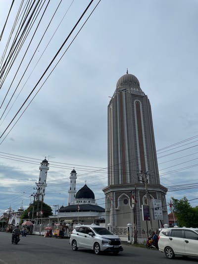 The image, originally rotated 90 degrees counter-clockwise, portrays a daytime street scene in Barat Yaman, Indonesia, under an overcast sky. A dense network of black overhead power lines crisscrosses the upper part of the frame.

To the right, a tall, grey, intricately designed minaret with a rounded dome and a textured surface dominates the scene. Adjacent to its base, a building displays several signs. One prominent sign advertises "LEMBAGA PENDIDIKAN DAN PENGEMBANGAN PROFESI 'ADHI GUNA'" (Institute for Education and Professional Development 'Adhi Guna'), mentioning "AKREDITASI B" (Accreditation B), "IJIN DIKNAS NO. 010.5/92/2012" (Education Department Permit), and "TERSEDIA BEASISWA 50%" (50% Scholarship Available). Below this, another sign shows a "YAMAHA" logo and the word "BAKSO."

Further down the street, a white mosque with a large, dark, patterned dome and two smaller white minarets is visible. Small Indonesian flags (red and white) are scattered along the street and buildings.

Street activity includes vehicles: a white car is prominent in the foreground, with its license plate "AG 1863 JY" partially visible. Another white car is parked near the tall minaret. A person on a red motorcycle, carrying yellow cargo, is seen driving past the tall minaret. The street is lined with other buildings, vehicles, and green foliage in the distance.