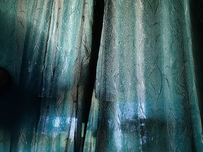 The image shows a close-up of textured teal curtains. The curtains have a raised, organic pattern that resembles leaves or vines. The lighting creates strong highlights and shadows, giving the fabric a three-dimensional appearance. The overall impression is one of depth and texture. The image is an interior shot, and it is not possible to determine its specific location within Ternovka, Moldova from the visual information provided.