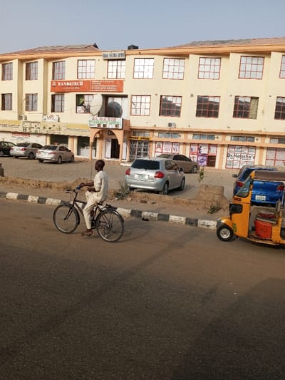 A daytime street scene captures an urban environment in Gyadi Gyadi Arewa, Nigeria. In the foreground, an asphalt road with a black and white striped curb is visible. A man, dressed in light-colored attire, is riding a dark-colored bicycle along the road, moving from left to right. To the right, a yellow auto-rickshaw (keke napep) is partially visible on the street.
Across the road, a dirt parking area hosts several parked cars, including a silver hatchback and other sedans. Behind the parking area stands a light yellow, two-story commercial building with brown trim and numerous multi-paned windows. A satellite dish is affixed to the building's facade.
Various business signs are displayed on the building. A prominent red banner advertises "RANDYTECH," listing services such as "MANAGED IT SERVICES," "NETWORK INFRASTRUCTURE," "IT CONSULTING & SERVICES," and "IT COMPLIANCE SERVICES." Other visible texts include a partial "EZA HAS AME" at the top, "LAFO," "NASIR" (likely part of "NASIR FURNITURE"), "AHMED NABIR," and "Dr. Natasha's." The clear sky indicates sunny weather, likely morning or late afternoon given the soft shadows. The scene depicts typical daily commerce and activity in a Nigerian urban setting.