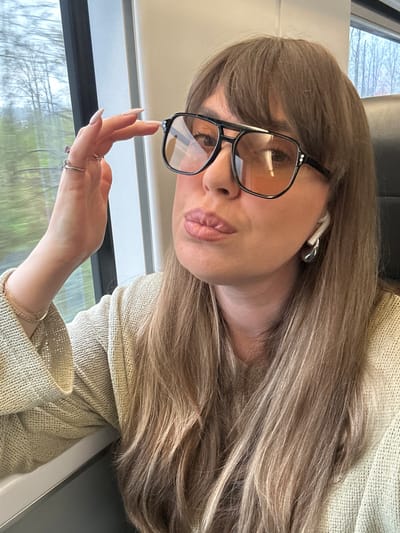 A woman with long brown hair and bangs is wearing eyeglasses and a tan sweater on what appears to be a train. She has an earbud in her left ear. She is holding her hand up to her face. The background shows trees through the train window, suggesting motion. There is not enough information to confirm the location is ქალაქი ლანჩხუთი, Georgia.