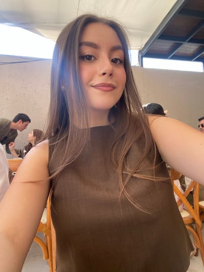 A selfie of a young woman with long brown hair, fair skin, and dark eyes. She is wearing a brown top. In the background, there is a man and another woman visible. They appear to be at a table. The location is Santa Cruz Tehuixpango, Mexico. Part of a canopy structure is visible in the top left corner. There are wooden chairs.