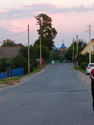 The image depicts a street scene in the village of Smilavichy, Smilavichy Sel'ski Sovet, Belarus. A paved road runs through the center of the frame, leading towards a cluster of buildings and trees in the distance. The sky above is a soft pink and purple, suggesting either dawn or dusk.
On the left side of the road, a blue fence borders a property with a house and trees. Several utility poles with wires are visible along the roadside. In the distance, a church with a blue dome and a golden cupola stands out against the trees.
On the right side of the road, there is a yellow building and more trees. A white car is parked on the side of the road, and a person is partially visible in the foreground on the right, wearing dark clothing with red accents. The overall impression is one of a quiet, rural setting.