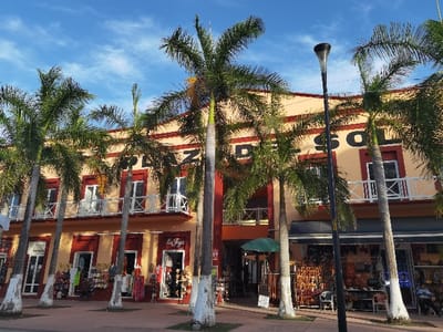 The image shows a row of shops in Cozumel, Mexico, located at latitude 20.51 and longitude -86.949. The shops are situated in a building with a peach-colored facade and a red trim, adorned with a black sign reading "Plaza del Sol". The building features several palm trees in front of it, and there is a black awning above the shops. The shops appear to be selling various goods, including leather items, clothing, and souvenirs. The street is lined with white painted curbs, and the shops have glass doors and windows. One person is visible in the image, a man standing outside a shop. The sky is blue with scattered white clouds. The sun is shining brightly, casting long shadows from the palm trees. There are two black lampposts on the sidewalk. The image is taken at an angle, providing a wide view of the shops and their surroundings.