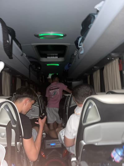 The image shows the interior of what appears to be a passenger airplane. Three young men are visible. One is seated, appearing to be looking at a phone. A second man in a pink shirt and grey shorts is partially visible in the middle of the aisle. The third man is seated and appears to be sleeping. Overhead compartments are visible along the length of the aircraft. Based on the data I have, I cannot confirm that it is taken in the city of Lesichovo, Bulgaria.