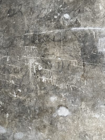 The image displays a close-up view of a textured, weathered concrete surface, likely a wall or floor. The surface is predominantly gray with variations in tone, indicating age and exposure. There are numerous markings on the concrete, appearing as scratches, lines, and smudges. Some of these markings are lighter, suggesting they were made with a hard object, possibly a sharp tool or by abrasion. There are also darker smudges, which could be dirt, grime, or old paint. In some areas, there are accumulations of white material, possibly plaster or chalk dust, which stand out against the darker concrete. The overall impression is one of an aged and possibly neglected surface bearing the marks of time and human activity. The location of Huejotzingo, Mexico, suggests this concrete surface could be part of historical architecture or an everyday urban structure. Without further context or visible features like signage or distinct architectural elements, it's impossible to specify the exact location within Huejotzingo.