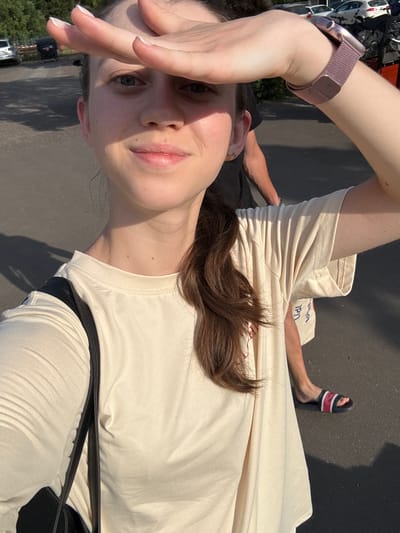 A woman is taking a selfie outdoors, holding her hand up to shield her eyes from the sun. She is wearing a light-colored t-shirt and a dark bag is visible over her shoulder. Her right arm, wearing a rose-gold colored smartwatch, is raised to her forehead. Her left leg is visible, wearing a black sandal with red and white stripes. Another person is partially visible in the background, wearing a dark t-shirt. The scene appears to be a paved area with some greenery and vehicles in the distance, suggesting a public outdoor space. The sunlight indicates it is daytime, likely a warm day. The text "LIDL" is visible on a white bag in the background. The image is oriented vertically.
