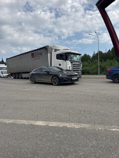 The image captures a scene on a paved road, possibly an intersection or a roadside rest area, in the city of Pesterevo, Russia. A large articulated truck, branded with "WEHA Trailer Group" and "SCANIA," is parked. In front of the truck, a dark-colored sedan, possibly a Kia, is also stopped, with its license plate visible as "H827XH 159." A white car is visible behind the dark sedan, suggesting a queue of vehicles. To the right, the rear of a blue car is visible. The environment appears to be outdoors with a cloudy sky overhead and a line of trees in the background. The road surface is asphalt with white markings. The image is taken from a low angle, possibly from within another vehicle.