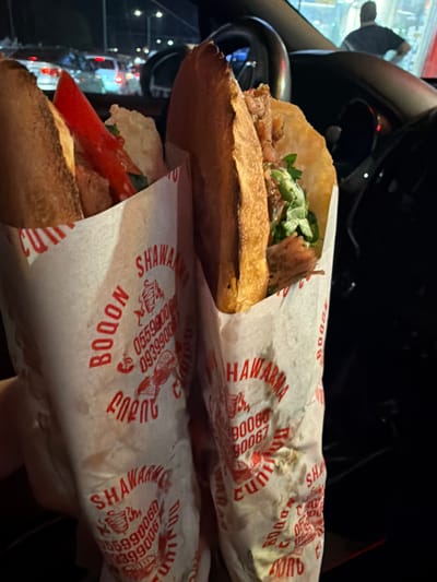 In the foreground, two shawarma wraps are held in white paper wrappers with red lettering. The wrappers display the words "SHAWARMA," "BOOON," and phone numbers such as "05599006," "093990067," and "09399006." One wrap contains meat, lettuce, and a red slice of pepper. The background shows a dark, blurry environment with car lights, suggesting a street scene at night. A silhouetted figure of a person is visible in the distance, standing near a vehicle. The setting appears to be a roadside, possibly in Parakar, Armenia, given the context.