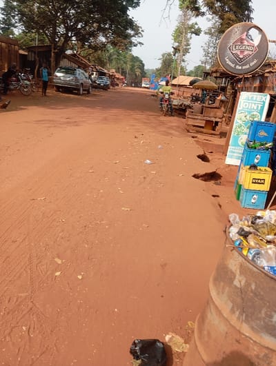 A dusty, reddish-brown road stretches through a rural African town. Vehicles and people dot the sides. On the right, a large "Legend" beer sign and "Fytex Joint" signage stand near stacked blue and yellow crates. A rusty metal barrel overflowing with trash sits in the foreground. Scattered litter is visible on the road. The overall impression is of a modest, bustling community with basic infrastructure.
