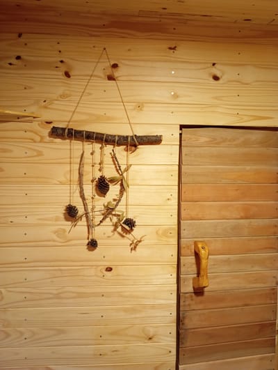 The image shows a wall made of light-colored wooden planks, with a sliding door made of darker wooden planks on the right side. A handmade mobile made of a branch, twigs, pinecones, and beads hangs on the wall. A rustic wooden handle is attached to the sliding door. The scene appears to be indoors, possibly in a cabin or a themed room. There are no people visible in the image. The lighting suggests it could be daytime or an interior light source is present. The image does not contain any identifiable landmarks or clues that would place it in Ensenada, Argentina.