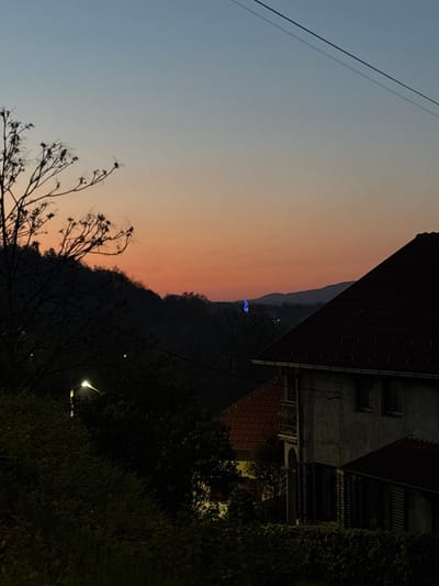 The image depicts a sunset scene in Vrnjacka Banja Municipality, Serbia. The sky transitions from blue to orange. A mountain range is visible in the middle ground. The silhouette of a building with a tiled roof and multiple windows occupies the upper right portion of the frame, partially obscured by vegetation. Bare tree branches are visible in the lower left corner. A streetlight is illuminated among the trees on the right. Powerlines cross the upper left sky.