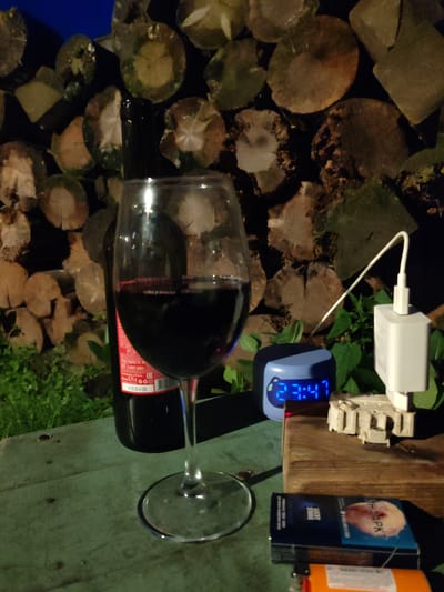 The image captures a nighttime scene outdoors, possibly in the city of Kashino, Russia. The foreground is dominated by a wooden table with a glass of red wine and a bottle of wine. Beside the glass is a digital alarm clock displaying "23:47" and a white USB charging adapter connected to a power source by a white cable. A pack of "LUCKY STRIKE" cigarettes and an orange lighter are also on the table. In the background, a pile of firewood is stacked behind the table, and green foliage is visible. The overall lighting suggests it is dark outside, with artificial light illuminating the immediate area.