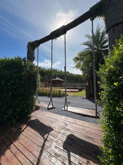 This image shows a swing set made of natural materials, including thick ropes and wooden beams. The swing set is set against a backdrop of lush green foliage and a bright blue sky with scattered clouds. In the distance, there are palm trees and a hint of a building with a thatched roof. The ground around the swing set is covered in dark gravel, and there's a wooden deck to the right of the frame. The sun is shining brightly, casting long shadows on the wooden deck, suggesting it is daytime, possibly late morning or early afternoon. The scene appears to be set in a tropical or subtropical location. There is no visible activity or people in the image, giving a sense of tranquility and a quiet, natural setting. A small sign with an icon is visible on a wooden post in the background, but the text is not legible. The overall impression is one of a peaceful outdoor space, perhaps a park or a resort, in Thailand.