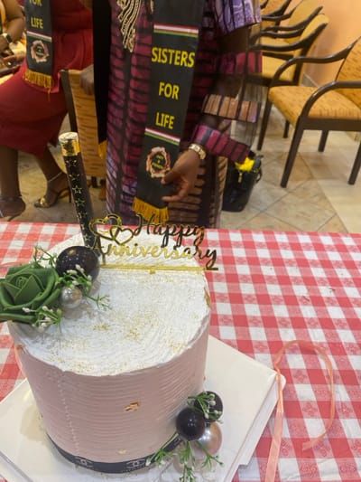 The image shows a cake on a table with a red and white checkered tablecloth in a location within the city of Buea, Cameroon, with the coordinates 4.145 latitude and 9.23 longitude. The cake is decorated with a green succulent, black and white round decorations, and a gold "Happy Anniversary" topper. A pink ribbon sits on the table next to the cake. A woman in a colorful, long-sleeved dress is visible in the background holding a black sash with the words "Sisters for Life" printed on it. She is holding the sash in front of her body with her right hand, while her left hand is on her hip. The back of a red dress is visible on the left side of the image. The scene is indoors, likely in a restaurant or a similar venue. The floor is tiled and the walls are painted a light beige color.