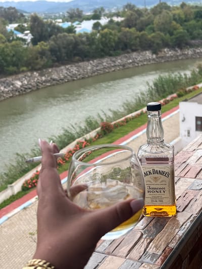 The image captures a scene of leisure and indulgence. A bottle of Jack Daniel's Tennessee Honey whiskey and a glass filled with ice and what appears to be the whiskey are placed on a tiled surface. A hand, adorned with a gold bracelet and manicured nails, is holding a lit cigarette.
In the background, a canal or narrow body of water runs parallel to a walkway lined with greenery and red flowers. Beyond the water, a cityscape can be seen in the distance. Based on the location in the city of Fnideq ⴼⵏⵉﺩﺍⵇ الفنيدق, Morocco, the scene is likely set on a terrace or balcony overlooking a waterway, possibly connected to the Mediterranean Sea or a nearby lagoon.