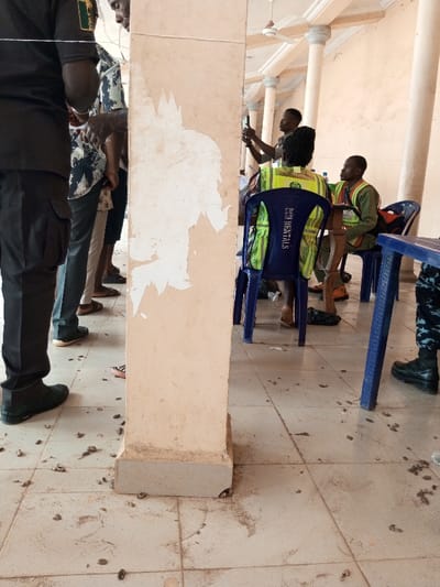 The scene depicts an indoor area within a building in Ajalli, Nigeria, characterized by a light-colored tiled floor and structural pillars. A prominent square pillar in the foreground, center-left, shows extensive peeling beige paint, revealing a lighter substrate. The tiled floor is visibly soiled with numerous small, dark, irregularly shaped pieces of organic debris and dirt.
On the far left, a person is partially visible, wearing dark pants and black boots. To the immediate left of the central pillar, several individuals are standing, appearing to form a line; their lower bodies are visible, dressed in various casual pants and footwear including sandals.
On the right side of the image, two individuals are seated on blue plastic chairs. The person closest to the camera is seen from behind, wearing a bright green and yellow reflective vest with "RENTALS" printed vertically on the back. To their right, another individual sits facing left, also wearing a similar green and yellow reflective vest. Behind these seated individuals, a person stands, holding a black smartphone horizontally at eye level, seemingly recording or photographing. A blue plastic table is partially visible on the far right.
In the background, multiple round, light-colored architectural pillars are visible, supporting a ceiling where a white ceiling fan is mounted. The overall lighting suggests daytime.