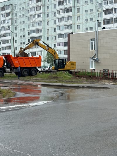A construction scene unfolds on a cloudy day in Usinsk, Russia, with a large orange dump truck positioned next to a yellow excavator. The excavator's arm is lowered, with its bucket holding dirt, suggesting it's in the process of loading or unloading material. The dump truck appears to be waiting to receive or transport the excavated earth. The ground is wet and muddy, indicating recent rain or damp conditions.

In the background, a multi-story apartment building with many windows dominates the scene, suggesting an urban or residential setting. The building's facade shows some signs of wear or aging. A metal pipe extends from the building, likely part of its infrastructure. A temporary construction barrier with red and white poles is visible along the edge of the work area. A white vehicle, possibly a van, is parked behind the excavator.

One person, wearing a dark blue jacket, is visible near the dump truck, potentially overseeing the operation or preparing to load. The overall atmosphere suggests ongoing construction or renovation work. The wet pavement and overcast sky point to a potentially cool and damp day. The visible text on the dump truck includes "116" and "NEFAZ."