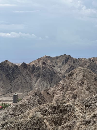 The image shows a mountainous landscape in Jiayuguan, China, with a prominent pagoda and a smaller pavilion visible. The foreground consists of rugged, rocky terrain with sparse vegetation. In the middle ground, a multi-tiered pagoda with a green tiled roof stands on a small hill. Further back, a pavilion with a traditional roof is situated on a hilltop. The background features a series of undulating mountains under a partly cloudy sky. The overall impression is one of a remote and historical site within a natural setting.