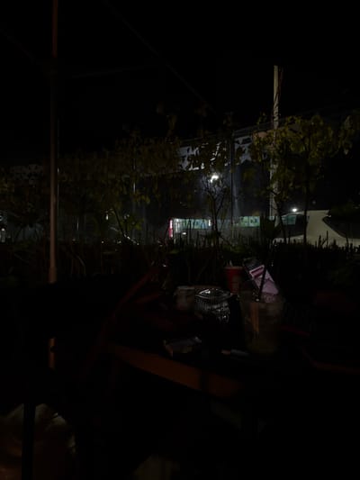 A nighttime shot reveals a blurred table with various items, including a clear plastic bottle, a can, and a pack of cigarettes. Outdoor vegetation frames the scene with glimpses of distant buildings and streetlights, creating a depth perspective. The overall ambiance is dark, with the objects on the table being partially illuminated and blurry. The image has been rotated and needs to be viewed sideways.