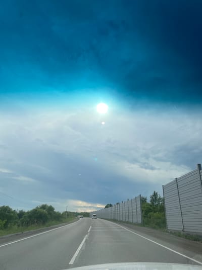 An overcast sky with a bright sun peeking through dominates the upper portion of the image. The sun is a bright white orb with a slight lens flare. Below the sky, a road stretches forward, curving slightly to the left. On the right side of the road, a tall, gray sound barrier runs along the highway. The barrier is made of vertical, corrugated panels. To the left of the road, dense green trees and foliage line the roadside. Several cars are visible on the road, moving away from the viewer. The scene appears to be a highway in or near Sosnovka, Russia, judging by the provided context.