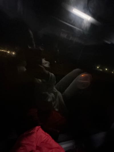 The photo depicts an indistinct scene in very low light, likely inside a vehicle or enclosed space. A long light fixture is visible in the upper left. There's a person under a white blanket or cloth and a red blanket on the right. Other details are obscured by the darkness, making it difficult to discern specific shapes or objects.