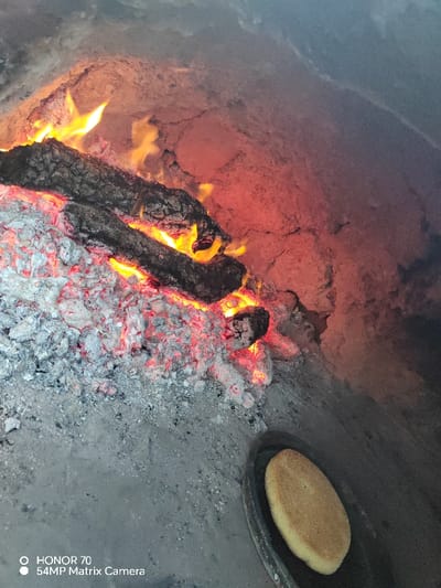 The image captures an inside view of a rustic, wood-fired oven, likely made of clay or brick, filled with glowing embers and active flames. On the left side, several dark, partially burnt logs are visible, interspersed with bright orange and yellow flames and a large bed of grey and red-hot embers and ash. The heat from the fire casts a vibrant reddish-orange glow onto the textured, rough inner walls of the oven, particularly prominent in the upper right and central areas, suggesting significant heat retention.

The floor of the oven, stretching towards the foreground and bottom right, is covered with a layer of fine, grey ash. In the bottom right corner of the frame, a single, round, flat piece of light-colored dough or unleavened bread is placed on a dark, oval-shaped pan or baking surface. The bread appears golden or light brown, indicating it is either partially baked or just starting to cook in the intense heat of the oven.

The overall scene conveys an atmosphere of traditional cooking or baking. The lighting is dominated by the warm, flickering light of the fire. In the bottom left corner, there is a visible watermark indicating the camera used: "HONOR 70" and "54MP Matrix Camera".