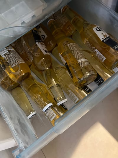 This media file shows a close-up view of a clear plastic container filled with multiple bottles of Corona beer. The bottles are predominantly light yellow in color, with distinctive white and black labels. The labels display the Corona logo, along with text in various languages, including "CERVEZA," "BRASSEE SOUS LICENCE," and "33cl/330ml." Some bottles have visible bubbles, suggesting they are cold. There are also some translucent, ice-like cubes visible in the container, likely to keep the beer chilled. The lighting suggests an indoor setting, possibly a refrigerator or cooler. The perspective is from above, looking down into the container. The overall scene is one of refreshment and leisure. The visible text indicates the brand and volume of the beer.