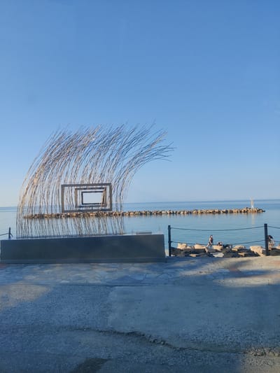 An outdoor scene captures a metallic sculpture with a wave-like structure comprised of thin, vertical rods and rectangular frames, situated on a concrete platform facing the sea. Beyond the sculpture, a breakwater constructed of large rocks extends into the calm, blue water. On the breakwater, a small white lighthouse stands to the right. In the foreground, a paved area leads to a beach with scattered seating cushions, where three individuals are visible: one person sitting on a cushion, another lying down, and a third person standing near a railing. The sky is clear and blue, indicating fair weather. The overall setting appears to be a seaside promenade or park in Platamonas, Greece, likely during daylight hours.