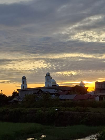 This image captures a sunset or sunrise over Keude Lueng Putu, Indonesia, focusing on a prominent mosque against a dramatic sky. The upper two-thirds of the frame are filled with a complex cloud formation, featuring dark grey and lighter grey clouds arranged in textured layers. Near the horizon, particularly across the middle and right sections, the sky transitions to intense golden-orange and yellow, indicating the obscured sun's position. This warm light illuminates the undersides of the clouds, creating horizontal streaks of color.

In the midground, a mosque stands centrally, distinguished by two white minarets with distinctive domed tops. A larger, lighter-colored dome is partially visible behind the right minaret. The mosque's main building appears to be a grey, multi-story structure. Surrounding the mosque are several other buildings, mostly in silhouette, with various roof designs. On the far right, a concrete structure reflects direct golden light. A tall, dark pole or antenna structure is visible on the far left, extending above the treeline.

The foreground features dense, dark green vegetation, including bushes and small trees, largely in shadow. In the lower mid-ground, a white bird is visible amidst the dark foliage. A narrow body of water, likely a canal or pond, is present in the bottom right corner, reflecting the warm hues of the sky. No individuals are discernible within the scene.