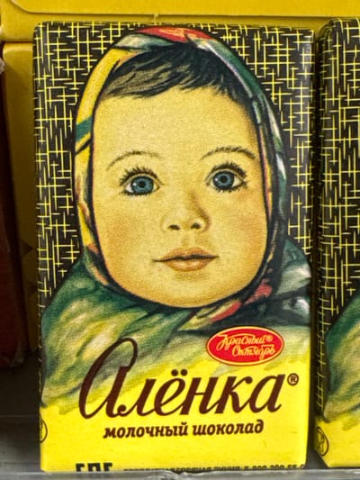 The media file shows a close-up, overhead view of a chocolate bar wrapper featuring a stylized illustration of a young girl's face wearing a headscarf. The girl has wide, blue eyes, a light complexion, and dark hair peeking out from under her headscarf. The wrapper is predominantly yellow with a black and yellow patterned border along the bottom.
Visible text on the wrapper includes the brand name "Алёнка" in large, stylized Cyrillic letters. Below this, smaller Cyrillic text reads "молочный шоколад," indicating "milk chocolate." To the left of the "Алёнка" text, within a red oval, is a logo with the text "Красный Октябрь," which translates to "Red October," a well-known Russian confectionery company. Another line of text is partially visible on the far right edge, appearing to be part of a product description or nutritional information.
The overall impression is one of a classic, perhaps nostalgic, product design. The illustration style suggests a traditional or vintage aesthetic. The setting appears to be a retail environment, possibly a shelf in a store, given the context of a product wrapper. No specific time of day, weather, or strong emotional cues are discernible beyond the innocent depiction of the child.