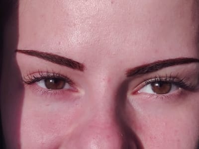 The image displays a close-up, front-facing view of a human face, specifically the area encompassing the eyes, eyebrows, and upper cheeks. Two open, brown eyes are centered in the frame, each featuring a visible pupil and white sclera. The eyelashes are long, dark, and appear volumized. Above each eye, a dark brown, well-defined eyebrow with a distinct arch is present. The skin across the forehead, around the eyes, and on the upper cheeks is fair-toned, exhibiting visible pores and a generally smooth texture with some minor surface variations. Direct, bright lighting illuminates the face from the right, casting a prominent, sharp vertical shadow along the left side of the face, partially obscuring the left temple and the outermost part of the left eyebrow. The subject maintains a neutral expression with a direct gaze. This scene is located in the city of Dolna Kremena, Bulgaria.