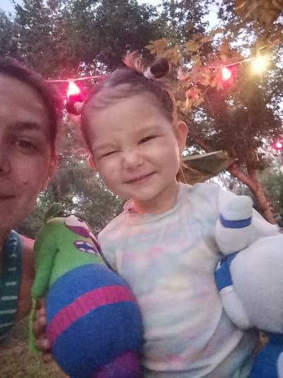 An outdoor, low-light, close-up selfie shows a woman and a young child with stuffed animals in front of trees and string lights. The woman, on the left, is mostly out of focus, with her left eye and cheek visible. She is wearing a green and white striped tank top. The child, in the center, is in focus, smiling with one eye closed and holding a green and blue stuffed animal with pink stripes and a purple section. Another white stuffed animal with blue accents is partially visible on the right. The background includes trees and illuminated string lights in red and yellow, suggesting an evening setting. The image is not identifiable to any specific location in Asprovalta, Greece.