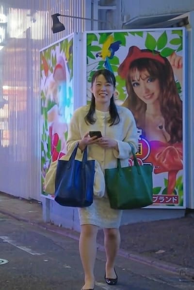 A Japanese woman stands on a city street in Gifu, Japan, smiling directly towards the viewer. She is holding a black smartphone in her right hand, a dark blue tote bag on her left arm, and a dark green tote bag on her right arm. She wears a light-colored, possibly cream, long-sleeved top or cardigan over a lighter dress or skirt, and black flat shoes. To her left is a light-colored, vertically ribbed architectural wall with a dark, articulated wall lamp extended from it. Behind her to the right are two illuminated advertising panels. The panel immediately to her right displays a large image of a smiling woman with long brown hair, wearing a red hat and a low-cut top that reveals cleavage, with a prominent heart-shaped decoration or tattoo visible on her chest. A blue and yellow parrot is depicted flying in the upper left of this advertisement. Japanese text, including "ブランド" (brand), is visible in the lower right portion of this panel. The street surface is dark pavement.