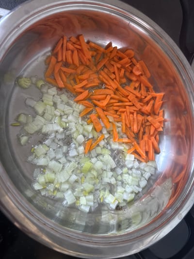 A stainless steel pot is on a black stovetop. Inside the pot, chopped carrots and leeks are being sauteed in oil. The carrots are cut into thin matchsticks and are a bright orange color. The leeks are chopped into small, irregular pieces and are white and light green. The oil in the pot is shimmering, indicating it is hot. The background is dark and out of focus. There is no visible indication of the city of Betzdorf, Germany.