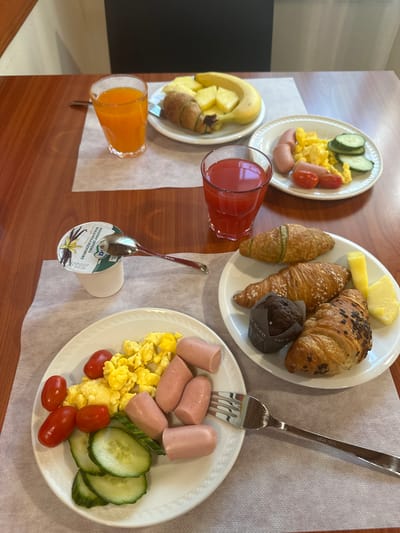 The image shows a breakfast spread on a wooden table, likely in a hotel or restaurant. There are three plates. The first plate (top left) contains scrambled eggs, sliced cucumbers, and halved cherry tomatoes. The second plate (center) contains a pat of butter, two croissants, and a muffin. The third plate (bottom right) contains sausages, scrambled eggs, sliced cucumbers, and halved cherry tomatoes. There is also a glass of orange juice, a glass of red juice, and a container of yogurt with a spoon. A fork is present on the rightmost plate. The image appears to be taken from an elevated perspective, looking down at the table.

The spoon shows the yogurt product is from Milk Spa, an Italian company. No other specific information is available on location in Villafranca di Verona, Italy, or on the identity of any people involved.