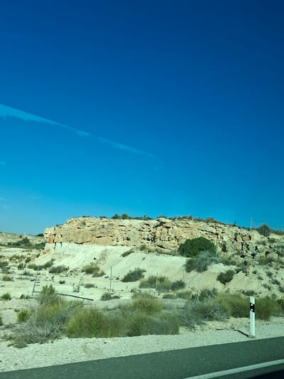 The image depicts a roadside view of a rocky, arid landscape under a clear blue sky. The foreground features a paved road with a white stripe, bordered by light-colored gravel and sparse, dry vegetation. In the midground, a low, rocky hill or cliff face is visible, with layers of light-colored rock and scattered green bushes. Some small, pale plants are also present on the slopes. A white traffic marker with black reflective bands stands on the right side of the road. The background shows a vast expanse of blue sky with a faint, thin white streak that may be a contrail. The scene appears to be in a dry, possibly desert or semi-arid environment. While the prompt mentions San Fulgencio, Spain, there are no specific visual cues in the image to confirm this location. There are no people or animals visible in the image, and no discernible actions or interactions.