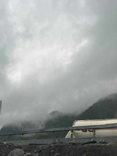 The image depicts a cloudy, overcast day in Røldal, Norway. The scene is dominated by a heavy, diffused cloud cover, suggesting a damp and cool atmosphere. In the foreground, a section of a white structure with red lettering is visible, partially obscured by a metal railing. The lettering appears to be "RØLDAL," possibly indicating a sign or the name of a building. Behind this structure, a steep, mist-shrouded mountainside rises, its upper reaches lost in the clouds. The overall mood is somber and atmospheric, characteristic of mountainous regions in overcast weather. There is no discernible activity taking place, and the focus is on the landscape and the man-made elements integrated within it.