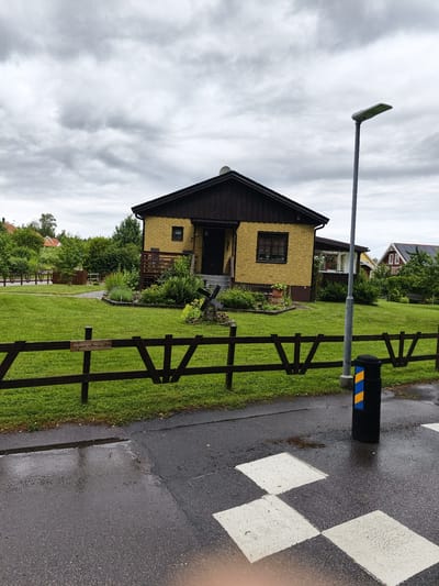 The image captures a street view in Ljungsbro, Sweden, on a cloudy day. In the foreground, a dark wooden fence runs horizontally across the frame, with a portion of a wet asphalt road featuring a crosswalk visible below it. A black cylindrical trash receptacle with blue and yellow markings stands to the right of the fence and road. Beyond the fence, a well-maintained green lawn leads to a yellow brick house with a dark brown roof and trim. The house has a front porch with a railing and a small window. To the left of the house, a garden area with various plants and a small decorative windmill is situated. In the background, several other houses and lush green trees and bushes are visible, indicating a residential neighborhood. The sky is overcast with heavy grey clouds. A street lamp is positioned near the house, to the right of the fence. A wooden sign is attached to the fence in the middle left of the image, bearing Swedish text.