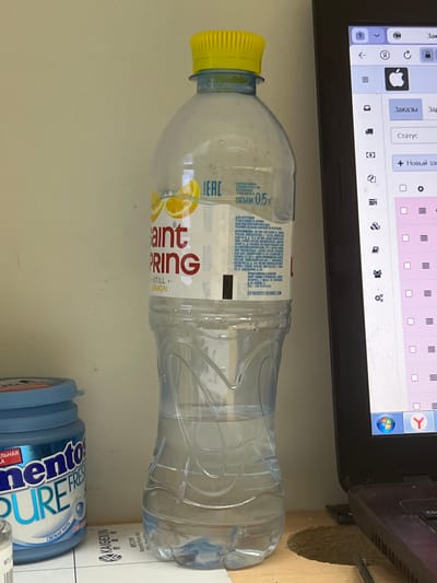The image shows a still life of everyday objects on a surface, likely a desk. In the foreground, a clear plastic bottle of Saint Spring still water with a yellow cap lies on its side. The label on the bottle is partially visible, featuring red text that says "aint PRING" and "STILL.. LEMON." Below the water bottle, a blue cylindrical container, possibly of Mentos gum, is visible, with "nento PURE FRE" and "СВЕЖАЯ МЯТА" (Fresh Mint) printed on it. In the background, a portion of a laptop screen displays various icons and text, suggesting it is powered on and in use. The overall setting appears to be indoors, with a neutral background and soft lighting, hinting at a daytime environment. No people or specific activities are depicted. The text suggests the location is Russia, with "Saint Spring" and "Mentos" being brand names.