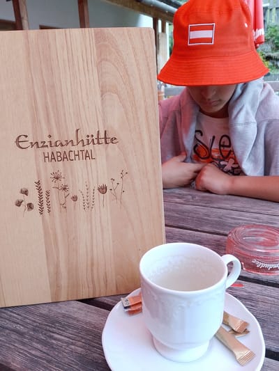 A close-up shot captures a wooden board with engraved text "Enzianhütte HABACHTAL" and decorative floral illustrations. To the right, a white teacup sits on a saucer, accompanied by two small packets of sugar. In the background, a person wearing an orange bucket hat with an Austrian flag emblem is visible, along with a grey hoodie. The setting appears to be an outdoor seating area, possibly a café or restaurant, with wooden tables and a rustic ambiance. The overall mood suggests a relaxed, casual moment, perhaps during daytime given the lighting. The visible text indicates the location as "Enzianhütte HABACHTAL" in Bramberg am Wildkogel, Austria.