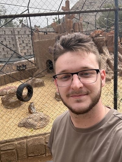 A male subject is depicted in a close-up photograph. He possesses short brown hair, a trimmed beard, and wears black-framed glasses and an olive-brown t-shirt. His expression is a slight smile. The subject is positioned in front of a wire mesh fence, behind which an animal enclosure is visible. The enclosure's ground is covered with light-colored organic material. A small monkey, exhibiting reddish-brown fur, is observed seated on a simulated rock formation in the mid-ground. Additional elements within the enclosure include a black tire positioned on a rock-like structure, and various artificial rock formations, one of which mimics a multi-level ancient building with numerous square apertures. Distant background elements include palm trees and an overcast sky. The setting is consistent with an animal exhibit within Jungle Park (Parque Las Águilas), located proximate to Los Cristianos, Spain.