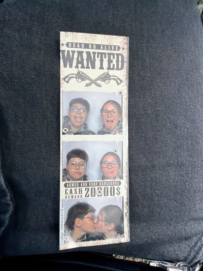The image is a vertical photo booth strip. The strip has a "DEAD OR ALIVE WANTED" theme. The top panel shows two people sticking their tongues out. The second panel also shows two people, one with a serious expression and the other with their tongue out. The third panel shows two people with surprised expressions. The bottom panel shows two people kissing. The photo strip has a code on the left side: "Codigo: FH9WAAXCCB". The background of the photo strip is a denim texture. The photo strip is placed on a dark denim fabric. There are no visible landmarks or indicators that place this image in the city of Sorø, Denmark.