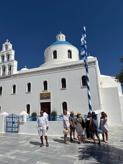 An eye-level, full shot captures a bright, sunny day in Ia Municipal Unit, Greece, with a whitewashed church as the central focus. The church features a prominent blue dome, multiple arched windows, and a bell tower on the left adorned with blue accents. In front of the church, a tall flagpole displays the Greek flag, striped blue and white, slightly unfurled.
A group of seven individuals stands in the foreground on a marble-tiled plaza. The person closest to the camera, a man wearing all white shorts and a button-down shirt with sunglasses, stands facing left with his hands in his pockets and a water bottle in his right hand. To his right, a group of six people, mostly women, are gathered, some wearing hats and summer attire, engaged in conversation. One woman in the group is wearing a blue dress and a wide-brimmed hat, her back to the camera, with her dress appearing to be caught by a breeze. The plaza is patterned with light and dark gray marble tiles, casting shadows from the building and people. Behind the group, a decorative blue and white fence borders the church's exterior. The sky is a clear, vibrant blue, and hints of olive tree branches are visible on the right side of the frame.