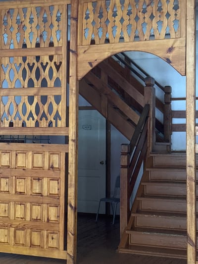 The image displays an interior scene within the city of Parizhskaya Kommuna, Russia, primarily composed of wooden architectural features. A light-toned wooden partition occupies the left and central-left foreground, featuring a lower section with multiple rectangular raised panels and an upper section showcasing repetitive, openwork carved designs consisting of curvilinear and pointed shapes. These sections are separated by a horizontal wooden beam. To the right, an arched wooden framework introduces a multi-step wooden staircase that ascends towards the upper right. The staircase is constructed from a darker wood, equipped with square balusters, and exhibits wear and chipped finish on its treads and risers. Directly behind and to the left of the staircase, a white door with a circular doorknob is visible against a light-colored wall. A small, dark rectangular sign bearing light-colored Cyrillic text is mounted above this door. A simple, light-colored metal-framed chair is partially visible in the background, positioned to the left of the door. The floor throughout the visible area appears to be dark wood. No individuals are present.